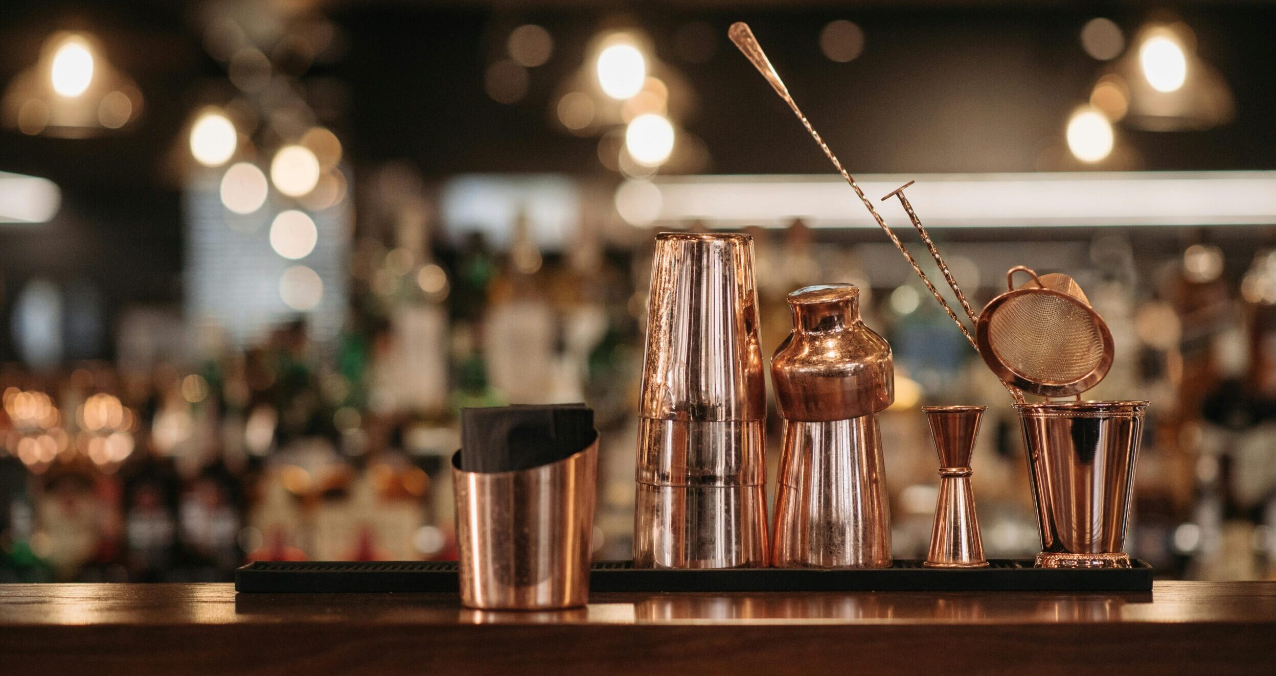 Elegant copper bar tools arranged neatly on a wooden bar counter with a blurred background.
